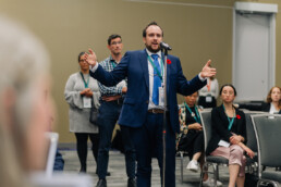Jeremy Roberts stands in front of a microphone stand with his arms outstretched as he asks a question. Behind him is a line of attendees waiting for their turn