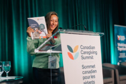 Emcee Katrina Prescott posing behind the podium with a copy of CCCE’s white paper, Giving Care on Day 2 of the Summit.