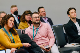 Audience members during a panel, with a bearded man wearing glasses in focus