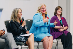 Laura Tamblyn Watts gestures with her hands and smiles during a panel on long term care. Seated on either side of her are Jodi Hall and Lisa Levin.