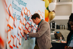 Un participant portant un costume marron et des lunettes épingle sa carte postale du Sommet sur un mur qui en est recouvert