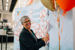 Dr. Zelda Freitas smiling as she pins her postcard to the Summit postcard wall. On the right is a bundle of orange and gold balloons