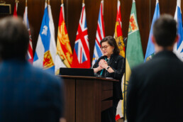 Dr. Naomi Azrieli giving a speech at the parliamentary reception with her hands clasped together. In the background is a collection of standing flags