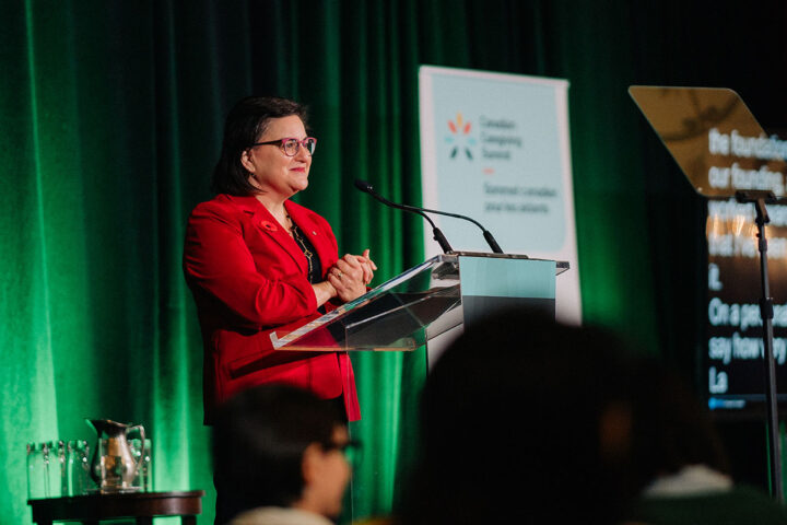 Azrieli Foundation’s CEO Naomi Azrieli stands at the podium with her hands clasped together.