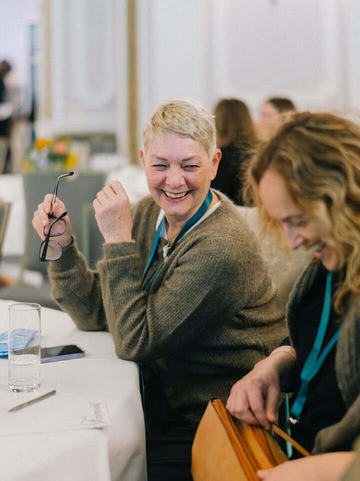 A woman with short blonde hair holds onto a pair of glasses with one hand while sharing a moment of laughter with her friend