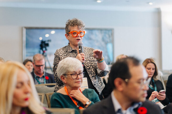 A woman with cropped curly grey hair and orange glasses stands up to ask a question during a session