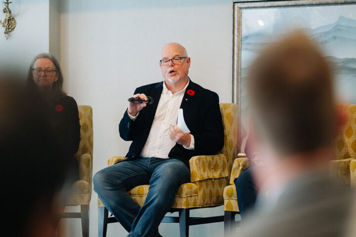 Colonel Russell Man seated in a yellow armchair, with the foreground showing blurred silhouettes of attendees