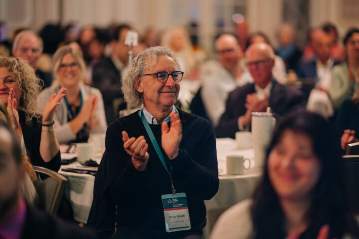 An older man with glasses and curly grey hair clapping his hands while surrounded by other attendees.