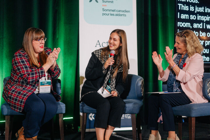 A young woman shares her advocacy idea on stage to applause from Amanda McKenzie and Kathleen Wynne, who are seated on either side of her.