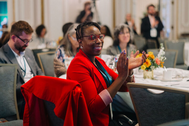 A smiling Black woman wearing a red blazer and red glasses applauds during the advocacy workshop