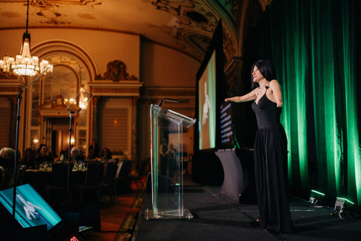 Side profile of comedian Courtney Gilmour on stage at the gala, with attendees seated in round tables on the left