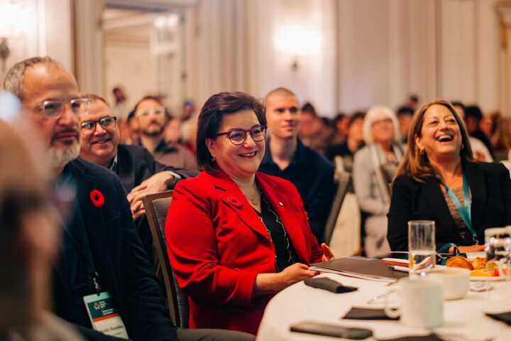 Azrieli Foundation’s CEO Naomi Azrieli smiling while seated at a table with other staff members. The background features other Summit attendees with their faces blurred.