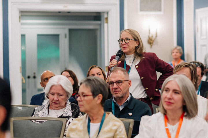 A woman with shoulder length blond hair and black glasses stands up to ask a question during a panel.