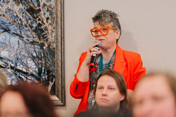 An audience member wearing an orange blazer, with short grey hair and orange glasses asks a question during a session.