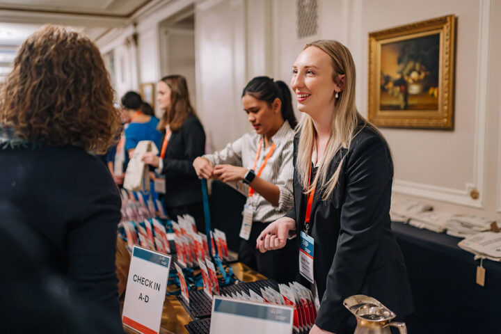 A smiling woman stands behind a check-in desk with badge holders at the conference assisting attendees with their registration.
