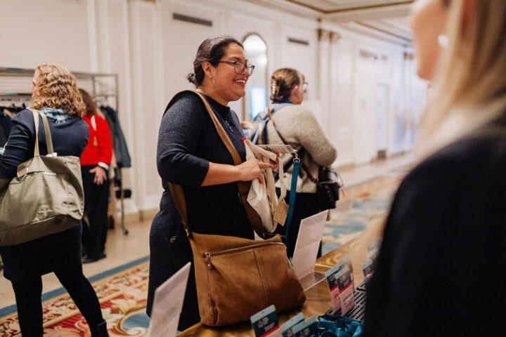 A woman with a brown bag and glasses smiles at the check-in desk while being helped. In the background are other attendees.