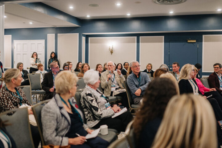 An older man with white hair and glasses asks a question during a panel while seated in a crowd of attendees