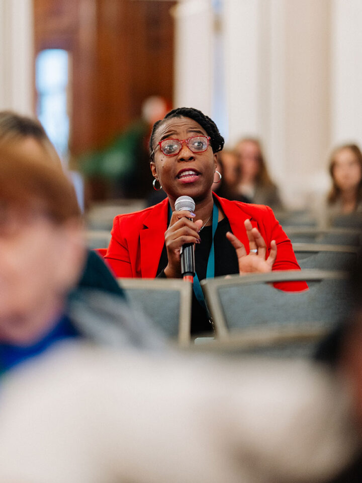 A Black woman wearing red glasses and a red blazer asks a question during a panel