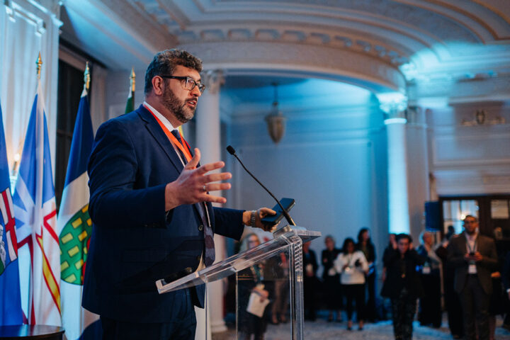 Side profile of CCCE's James Janeiro speaking on stage at the parliamentary reception, with attendees in the background.