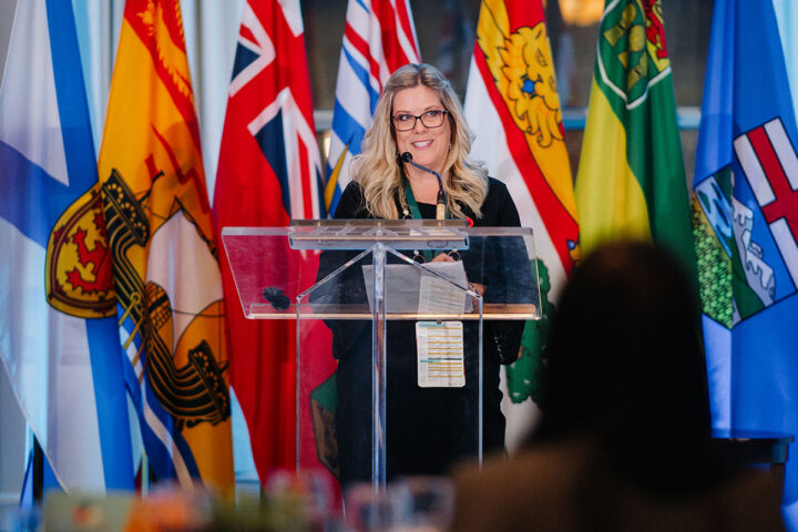 Brenda Blais, co-host of the parliamentary reception, speaks on stage against a background of hanging flags.