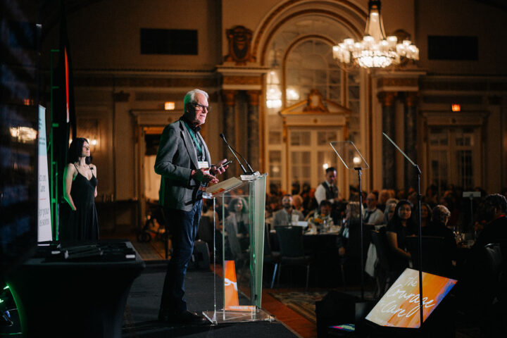 74. A side profile of Al Etmanski speaking on stage, with attendees seated in round tables below
