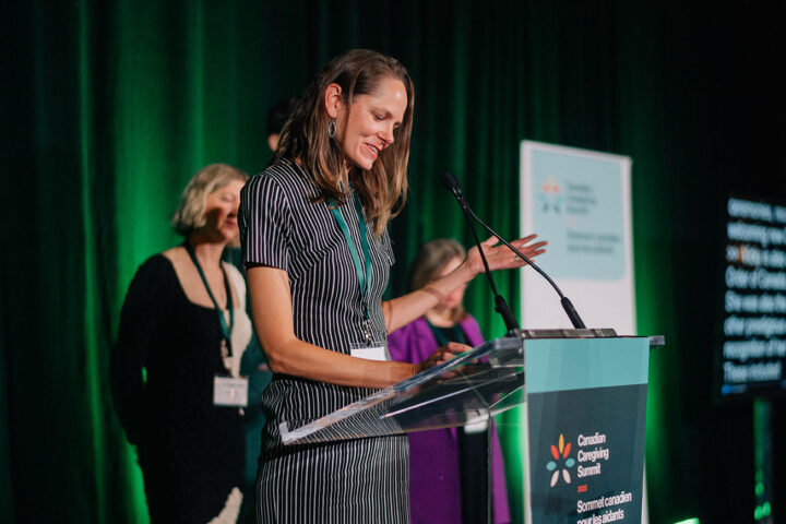 Catherine Etmanski speaks at the podium, with her siblings on stage in the background