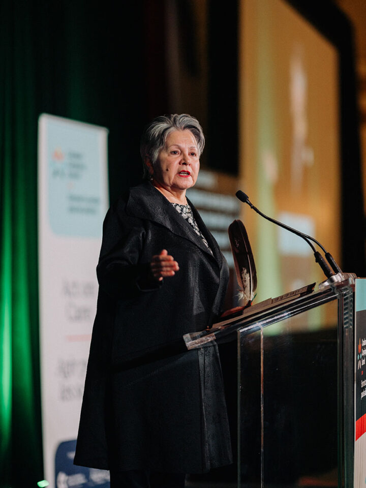 Elder Claudette Commanda speaking at the podium, with a feather grasped in her left hand.