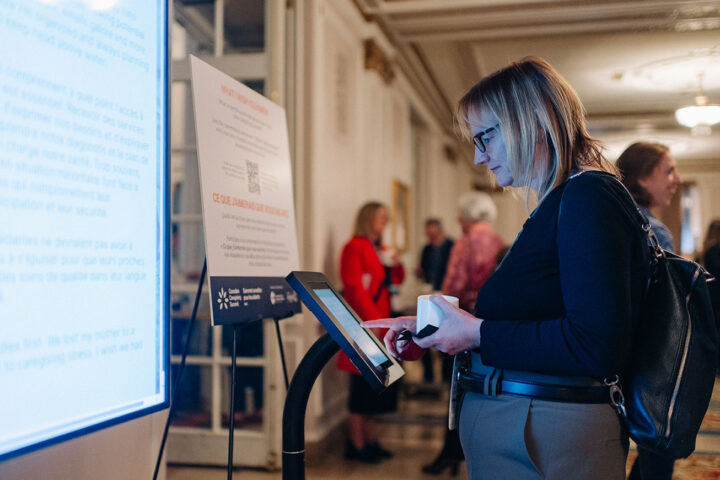 A woman with dark blonde hair and black glasses stands in front of an iPad and projector screen, where attendees can submit answers for the What I Wish You Knew activity