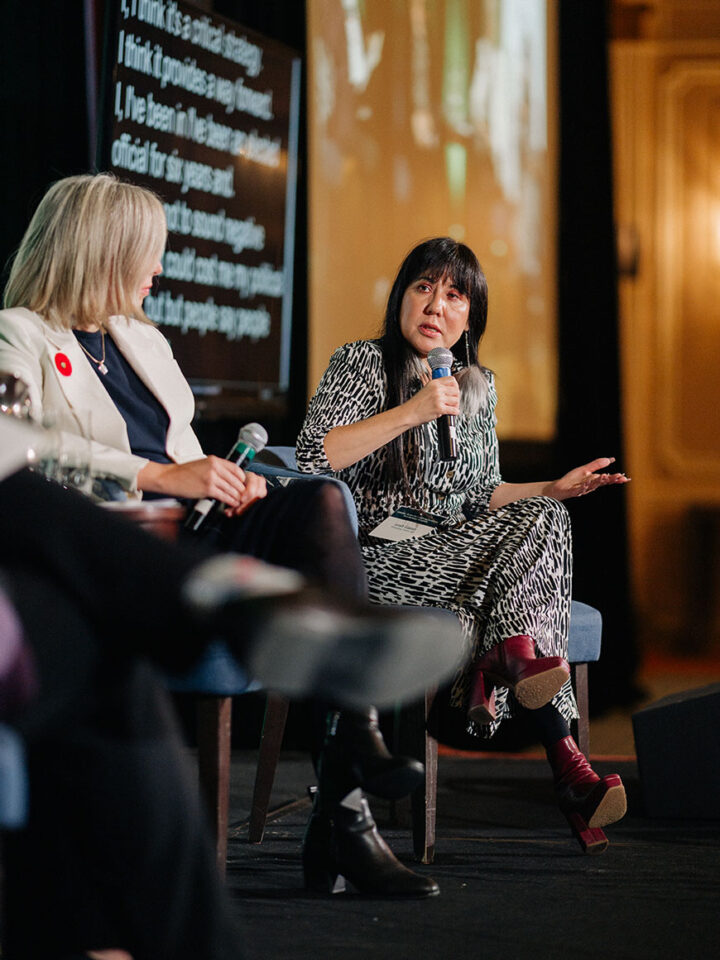 MP Leah Gazan speaks on stage, with MP Leslie Church seated next to her. Behind is a screen with subtitles.