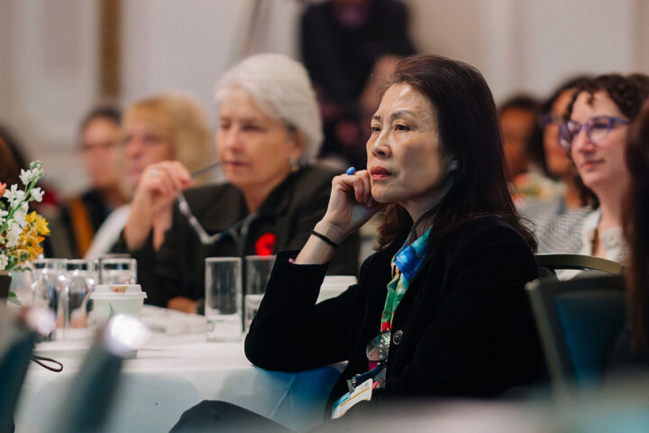 A middle-aged attendee with black hair sits with her head propped on one arm, with other attendees in the background.
