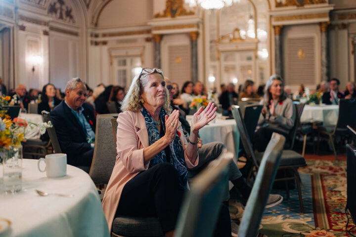 Former Ontario Premier Kathleen Wynne applauds while seated at a table. Behind her are round tables filled with attendees.
